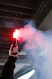 A close-up of a hand holding a smoking red flare under a bridge.